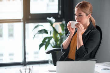 Poster - Portrait of pensive girl tasting cup of liquid while sitting at table in office. Job and rest concept. Copy space