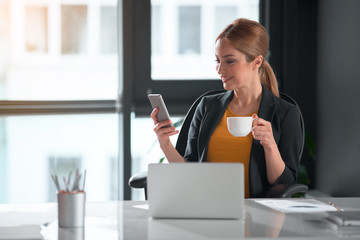 Poster - Happy businesswoman typing in phone while drinking mug of coffee in office. Break and job concept