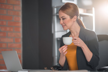 Poster - Side view beaming lady tasting cup of delicious tea while looking at screen of notebook computer in apartment. Relax concept