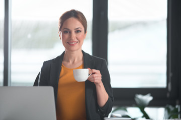 Poster - Portrait of happy businesswoman drinking mug of appetizing beverage while typing in laptop. Rest concept. Copy space