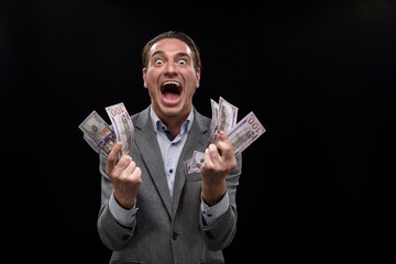 I am crazy about money. Portrait of young insane businessman is holding cash and looking at camera with wide-eyed. He is standing against dark background and screaming. Isolated