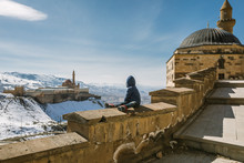Young Boy Sitting On A Stone Wall Looking To An Oriental Palace In Snowcovered Mountain Landscape, Turkey