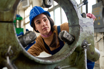 Portrait of young female mechanic wearing hardhat  looking into machine part while working at modern plant, copy space