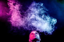 A Young Man Exhales A Cloud Of Colored Smoke Of Blue And Pink Color On A Black Isolated Color.  Guy Is Admiting Puffs Of Steam From The Electronic Cigarette.