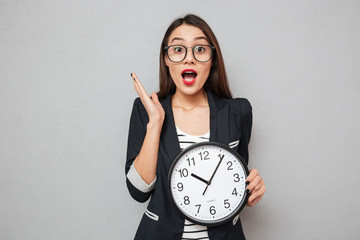 Poster - Surprised asian business woman in eyeglasses holding clock