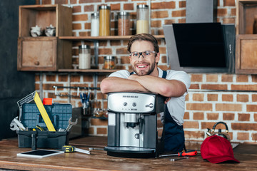 handsome young repairman leaning at coffee machine and smiling at camera