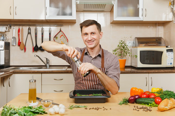 Wall Mural - Caucasian young man in apron sitting at table with vegetables, sprinkling pepper meat from mill, cooking at home preparing meat stake from pork, beef or lamb, in light kitchen with wooden surface.