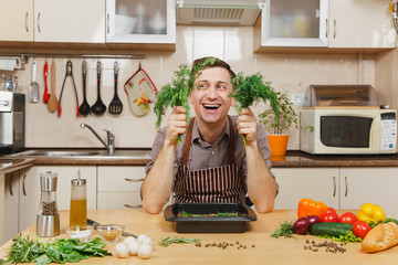 Wall Mural - Handsome fun young man in apron sitting at table with dill or fennel, cooking at home preparing meat stake from pork, beef or lamb, in light kitchen with wooden surface, full of fancy kitchenware.