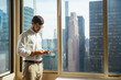 © AlejandroMCB photo & film/Stocksy - Young businessman in an office checking his phone with the view of city buildings on the background