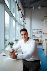 Young handsome businessman posing with tablet at office at a window . Short-haired clean-shaven man in white shirt looking so serious and demanding. Freelance. 