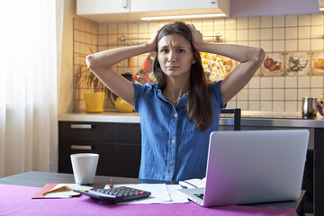 Upset uptight desperate housewife grabs his head sitting at the table in the kitchen because of financial problems. The concept of a home budget, loans, invoice, financial resources and sress.