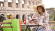 Beautiful young woman tourist reading menu sitting at the table of a bar restaurant in front of the Colosseum in Rome. Elegant dress with large hat and colorful shopping bags on a summer day.