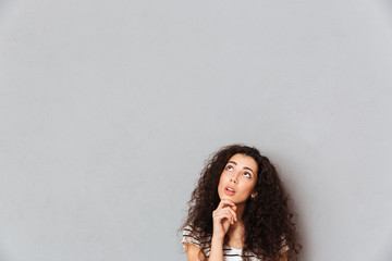 Poster - Concentrated young woman with shaggy hair touching her chin with face upward and thinking or dreaming over grey background in studio