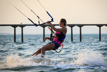 Wall Mural - Kite surfing girl in sexy swimsuit with kite in sky on kiteboard in the blue sea riding waves saying hi. Recreational activity, water sports, action, hobby and fun in summer time. Kiteboarding sunset