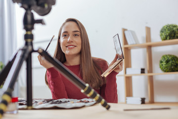 Fashion blogger. Cheerful pretty beauty blogger smiling to the camera and feeling glad while holding a brush and eye shadows