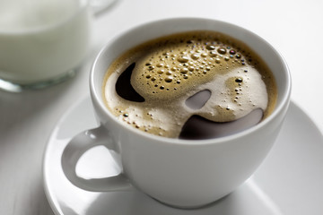 Freshly poured black coffee with froth in white ceramic cup next to a milk jug on white.