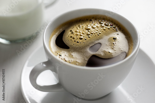 Fotografie, Obraz Freshly poured black coffee with froth in white ceramic cup next to a milk jug on white