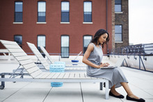Full Length Of Businesswoman Using Laptop While Sitting On Lounge Chair At Building Terrace