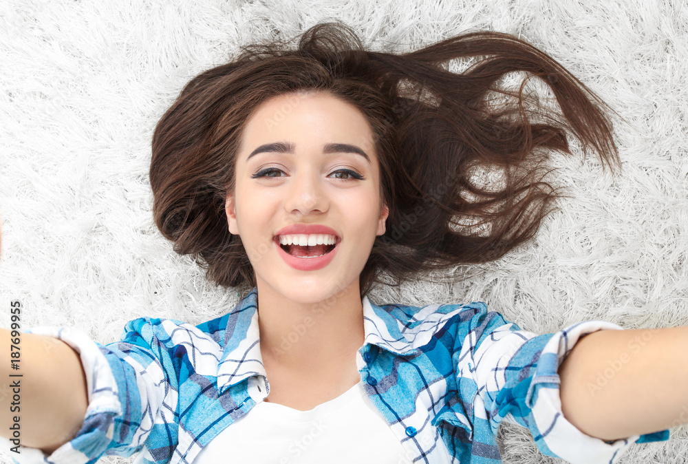 Beautiful smiling woman taking selfie while lying on carpet at home