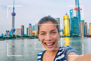 Wall Mural - Happy China travel selfie Asian tourist woman vlogging online recording videoblog vlog video. Smiling young girl holding camera phone at the Bund at Shanghai's skyline of skycrapers.