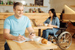 © zinkevych - Working process. Handsome cheerful well-built blond man holding his phone and writing in his notebook while a woman sitting in a wheelchair in the background