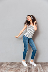 Wall Mural - Full size view of charming young woman in striped t-shirt and jeans dancing while listening melodies via headphones over grey background