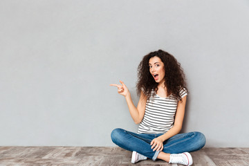 Poster - Lovely curly woman in casual clothes sitting in lotus pose on the floor pointing index finger aside submitting something over grey wall copy space