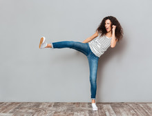 Portrait Of Strong Young Female With Curly Brown Hair Kicking Invisible Opponent Punching With Leg Over Grey Wall