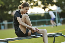 Teenage Girl Tying Shoelace While Sitting On Bench In Playing Field