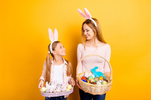 We Are Ready For The Celebration! Cute Lovely Nice Sweet Mommy And Small Daughter Holding Whisker Baskets With Colored Eggs  Cakes And Cookies Looking At Each Other Isolated On Yellow Background