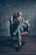 © deagreez - Cheerful, handsome, old, smiling boss in spectacules, suit watch, look, read newspaper, smoke cigarette, sitting in leather chair in workstation, wooden floor, gray background, relax, fun, fashion