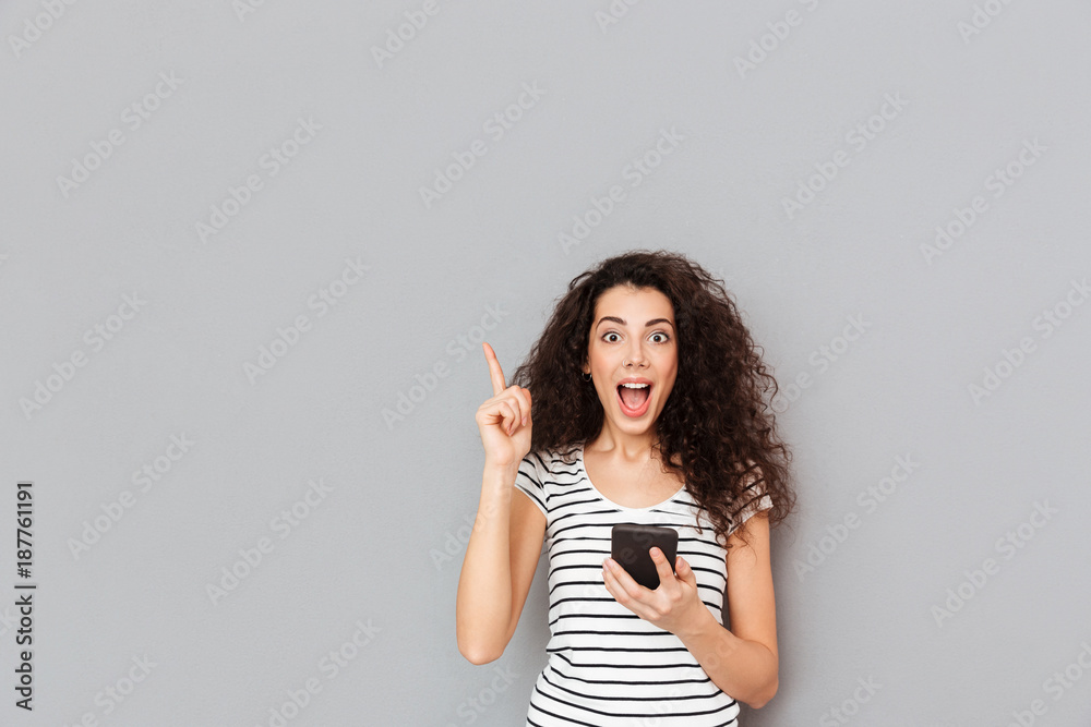 Smart female with mobile phone in hand keeping index finger pointed upwards expressing she has an idea or saying eureka over grey background - obrazy, fototapety, plakaty 