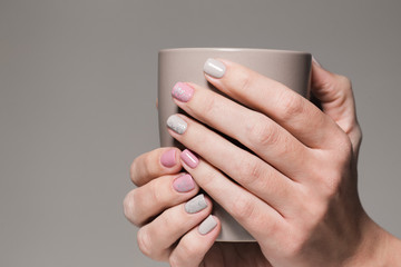 Closeup view of white hands of healthy woman with beautiful modern grey and pink manicure design. Woman holding brown cup. Horizontal color photography.