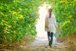 The girl walks along the path in the summer woods to the light in a white jacket and jeans.