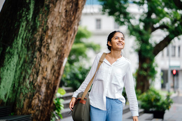 portrait of young smiling indian woman walking through the city wearing a bag. It is outdoors and there are trees and greenery in the background