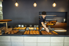 Man Working In A Bakery, Placing Freshly Baked Croissants And Cakes On Large Trays On A Counter.