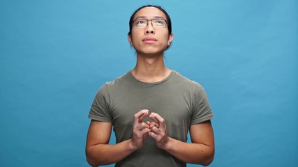 Poster - Pensive asian man in t-shirt and eyeglasses praying with crossed fingers over blue background
