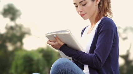 Sticker - Smiling brunette woman sitting on grass and writing something in notebook in park