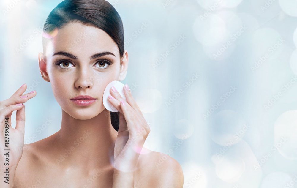 Young brunette girl removing makeup from her face. Photo of beautiful girl with cotton pads  on shiny silver background. Skin care and beauty concept. - obrazy, fototapety, plakaty 
