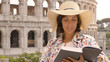 Young woman tourist reads a book sitting at the table outside a bar restaurant in front of the Colosseum in Rome with coffee, juice and cornetto. Elegant beautiful dress with large hat and colorful