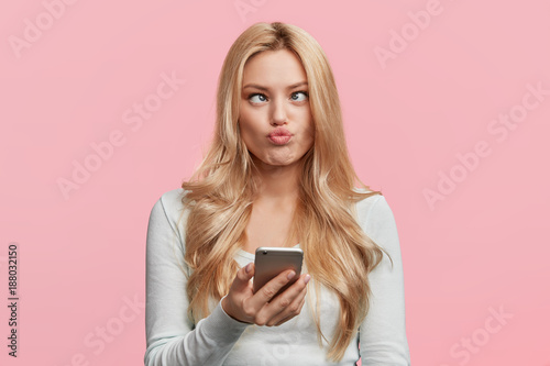 Cuadros en Lienzo Crazy funny comic female crosses eyes, pouts lips, makes grimace, foolishes, holds cell phone, involved in games online, isolated over pink background