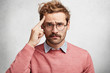 © WHstudio Leushin N - Portrait of handsome young stylish male tries to concentrate as sees something in front, has problematic expression, dressed in pink sweater, has round eyewear, isolated over white concrete wall