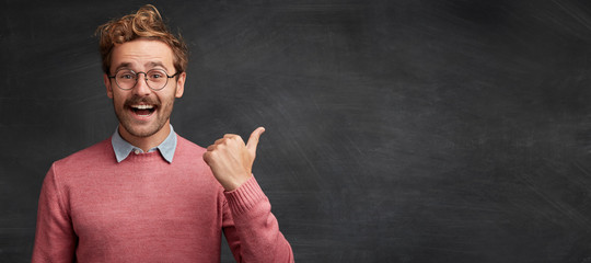 Poster - Horizontal shot of bearded fashionable male student indicates at blank chalk blackboard with thumb, has happy expression, advertizes something. People, positive emotions and promotion concept