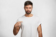 © wayhome.studio  - Serious pleasant looking bearded male brunet indicates at blank copy space of his t shirt, advertizes its quality, area for your design, logo or promotional text, isolated over white studio background