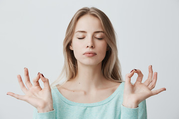 Wall Mural - People, yoga and healthy lifestyle. Gorgeous young blonde woman dressed in light blue sweater keeping eyes closed while meditating indoors, practicing peace of mind, keeping fingers in mudra gesture