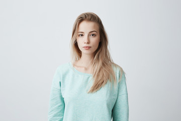 Wall Mural - People and lifestyle. Studio shot of attractive young Caucasian dark-eyed female with long dyed blonde hair posing against gray blank wall dressed in casual blue sweater with calm face expression.