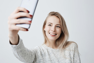 Wall Mural - Beautiful caucasian young female with long blonde dyed hair and appealing dark eyes holding mobile phone, posing for selfie, looking at camera with flirty smile. Attractive woman making video call.