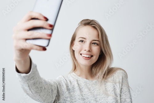 Fotomural Beautiful caucasian young female with long blonde dyed hair and appealing dark eyes holding mobile phone, posing for selfie, looking at camera with flirty smile