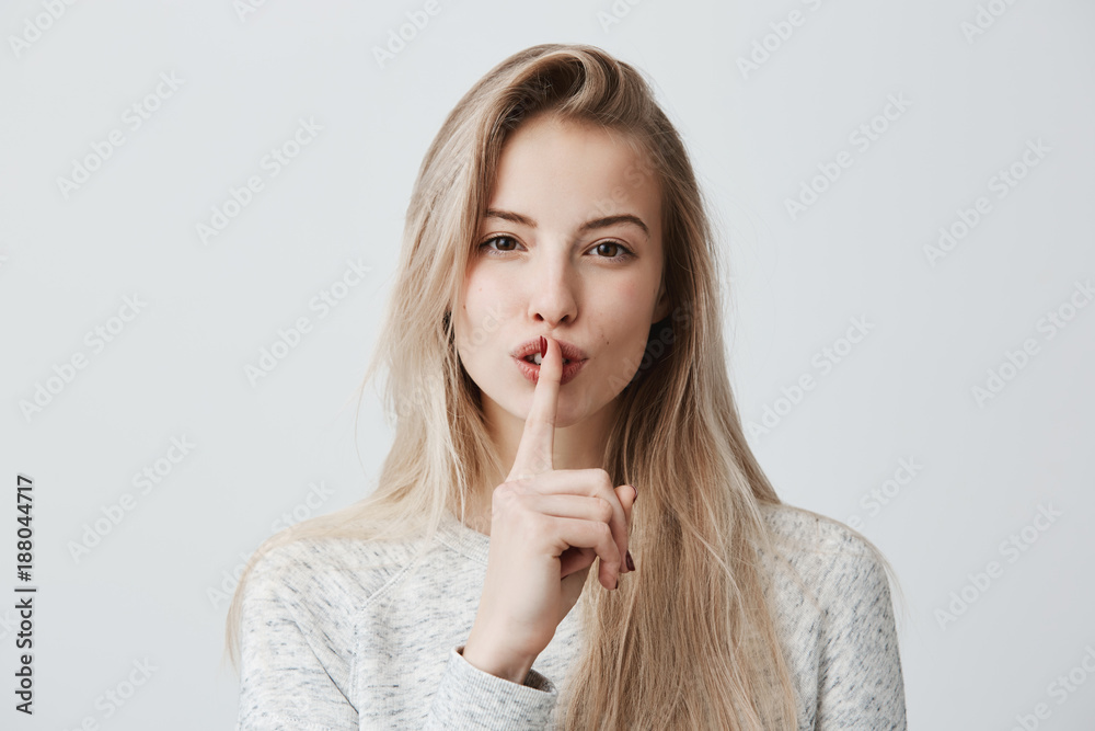 Shot of beautiful female with long blonde dyed hair wears casual long-sleeved t-shirt, shows hush sign, has serious expression, asks not to tell her secret anybody, hopes for loyality and silence. - obrazy, fototapety, plakaty 