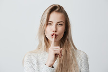 Shot Of Beautiful Female With Long Blonde Dyed Hair Wears Casual Long-sleeved T-shirt, Shows Hush Sign, Has Serious Expression, Asks Not To Tell Her Secret Anybody, Hopes For Loyality And Silence.